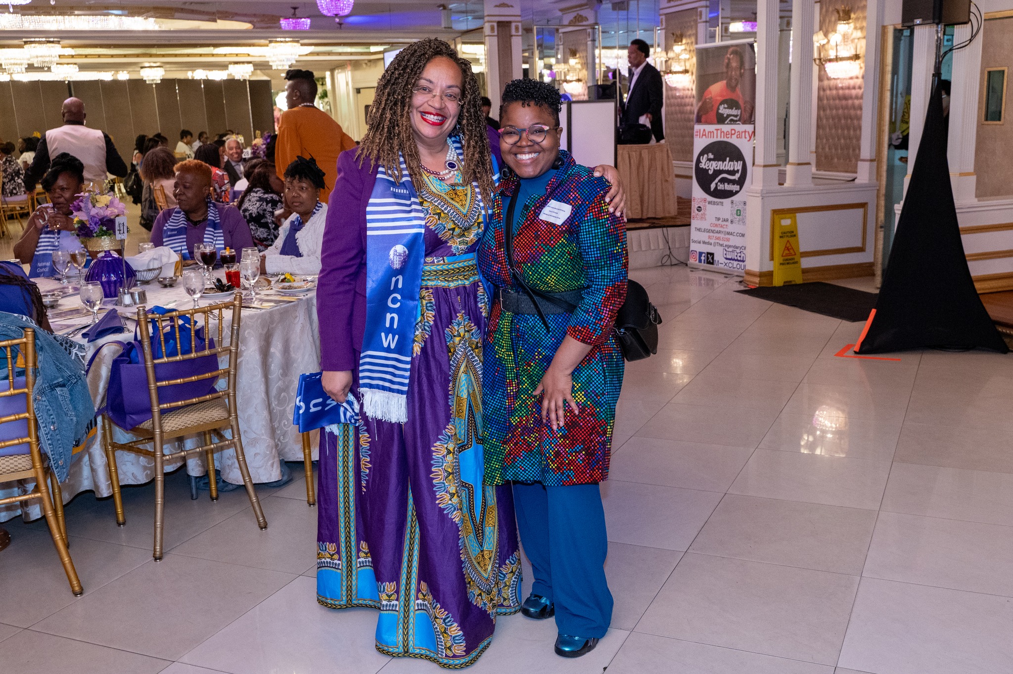 Women attending the New York State BHRP annual Luncheon in 2024. A photograph taken at an indoor event venue with two African American women standing together in front of a dining table. The woman on the left wears a vibrant purple and blue African print dress with intricate patterns and a matching blue scarf with "NCNW" text. The woman on the right wears a blue and red patterned outfit with a blue headscarf. Both are smiling warmly at the camera. Behind them is a formal dining table with gold Chiavari chairs, white tablecloths, and wine glasses. The venue has white tile flooring and decorative lighting fixtures on the ceiling. In the background, there are other attendees seated at similar tables. A black banner is partially visible on the right edge of the frame. The lighting is warm and even throughout the space, creating a formal event atmosphere.