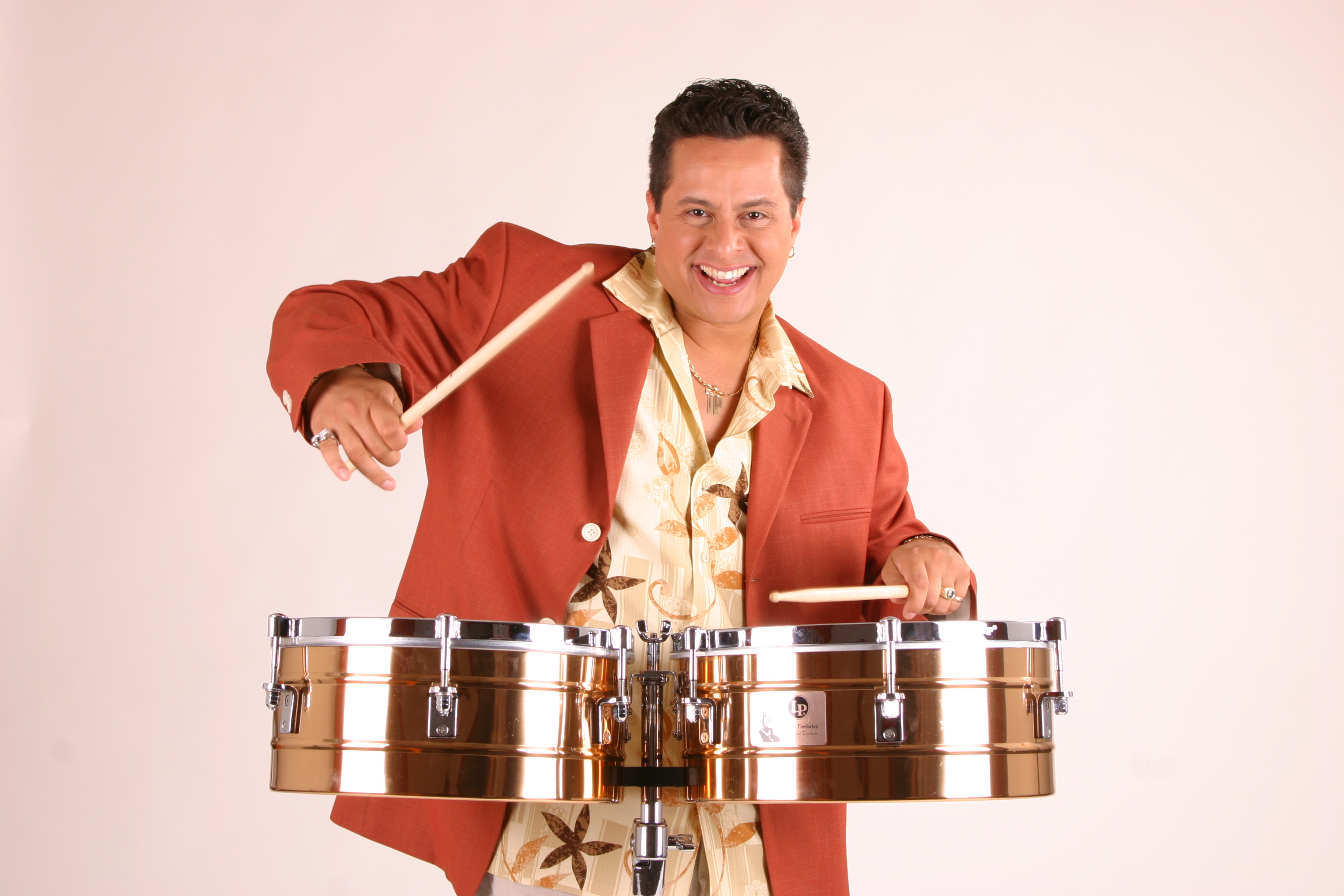 Tito Puente Jr. performing on timbales with drumsticks and a bright smile.