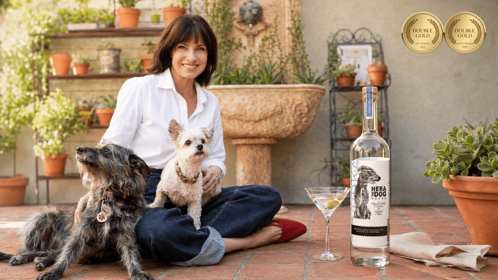 Julia Pennington sits with two dogs beside a bottle of Hera The Dog Vodka, a martini glass, and Double Gold award medallions