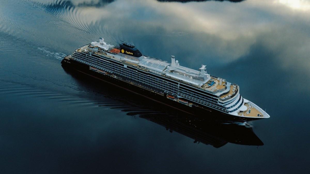 Aerial view of the EXPLORA III luxury cruise ship sailing across calm, dark water that reflects the clouds above