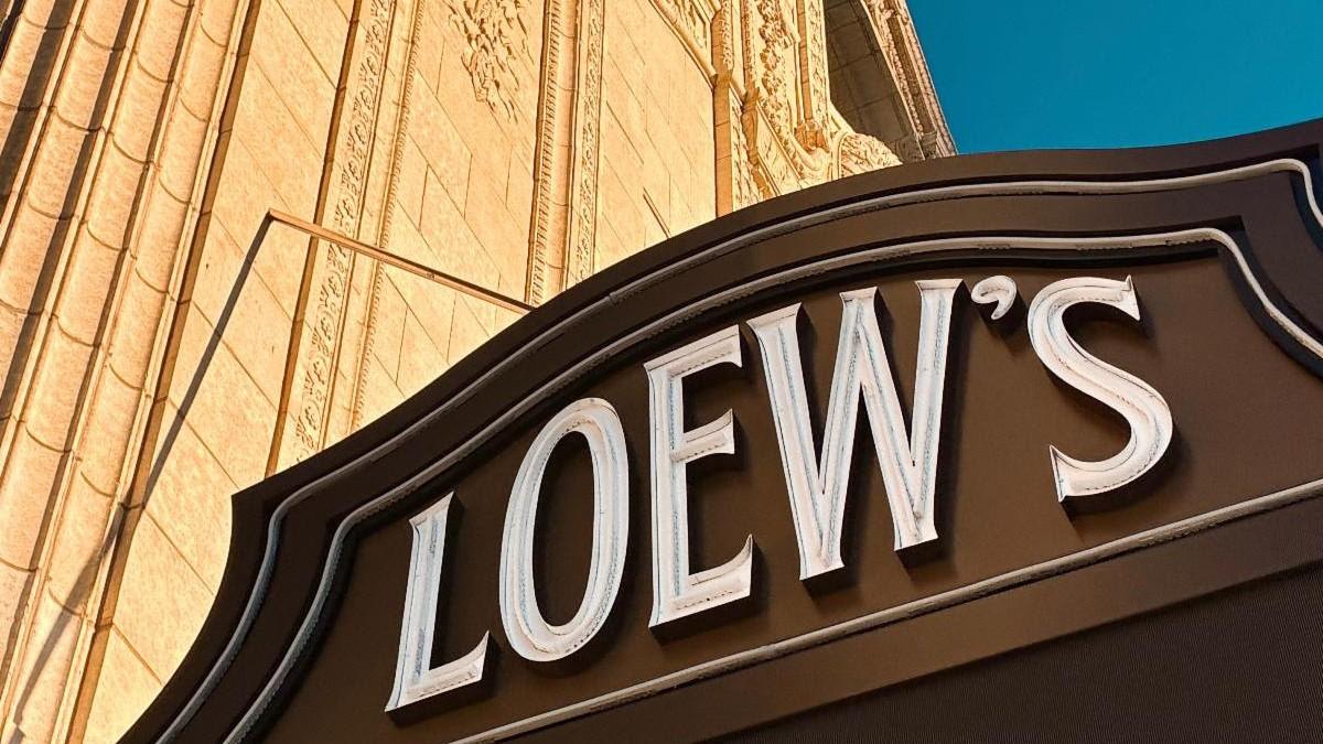 Restored Loew’s marquee with white lettering on a brown sign beneath the theatre’s ornate golden terra-cotta facade and blue sky