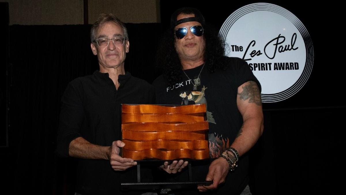 SLASH in mirrored sunglasses holding a carved copper-toned Les Paul Spirit Award beside a presenter, with the award logo behind them